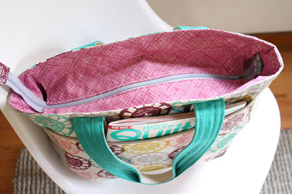 A fabric tote bag with a pink patterned interior and teal handles, shown resting on a white chair. The exterior of the bag has a multicolor floral print on a cream background.
