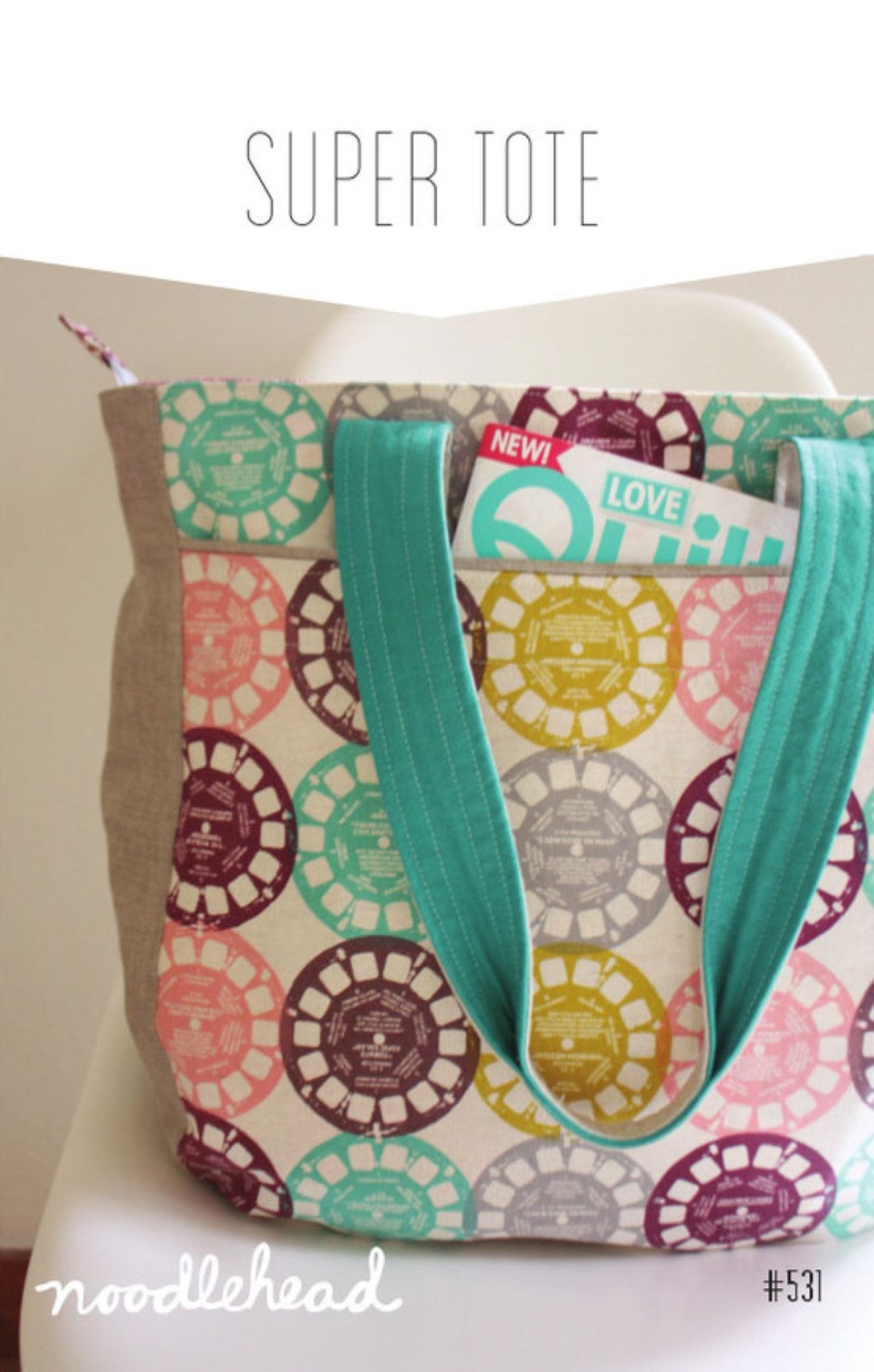 A tote bag with a colorful circular pattern in pink, teal, yellow, and purple on a cream background, featuring solid teal fabric handles and a natural beige side panel. The bag is shown sitting on a white chair with text overlays reading 'SUPER TOTE' and 'noodlehead'.