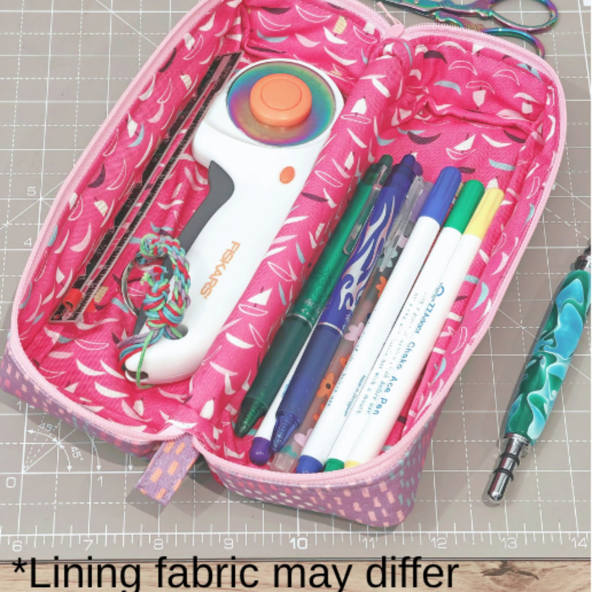 An open pink patterned fabric zip pouch or pencil case containing a rotary cutter, pens, and other sewing/craft tools, photographed on a gridded cutting mat with a text note at the bottom saying the lining fabric may differ.