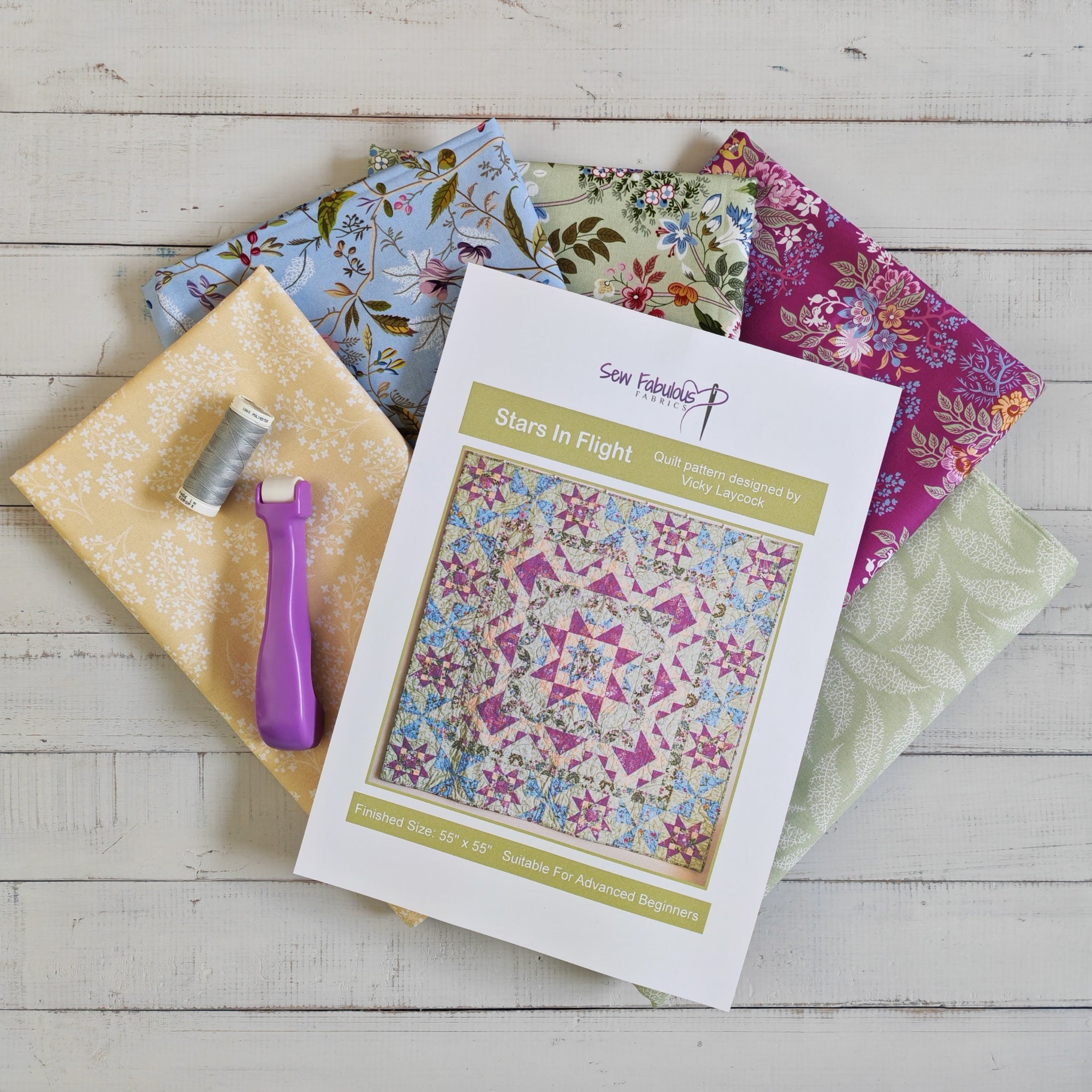 A quilt pattern displayed on top of several folded patterned fabrics, with a purple seam roller and a spool of thread, arranged on a whitewashed wooden surface.
