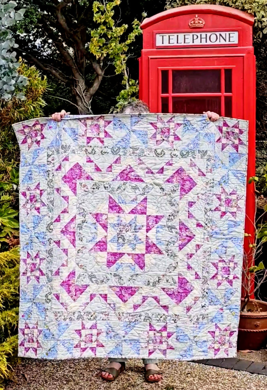 A person holds up a quilt featuring pink, purple, and blue starburst patterns on a white background, set against a garden backdrop with a red telephone booth.