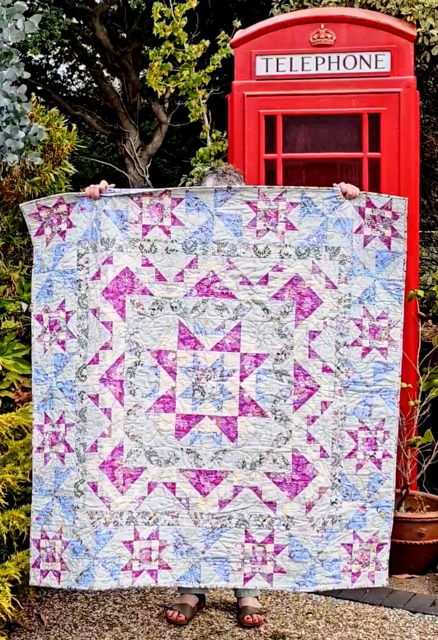 A person holds up a quilt featuring pink, purple, and blue starburst patterns on a white background, set against a garden backdrop with a red telephone booth.