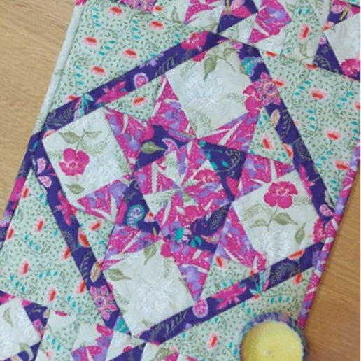 A colorful patchwork quilted table runner with floral and geometric patterns, displayed on a wooden surface next to a small yellow candle.