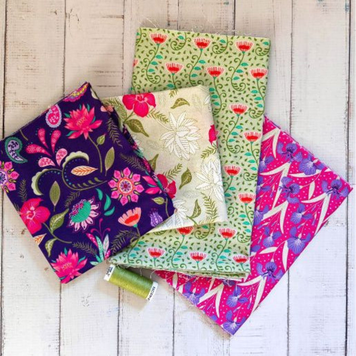Four folded fabric pieces with colorful floral and paisley patterns arranged on a white wooden surface, with a small spool of green thread resting near the center.